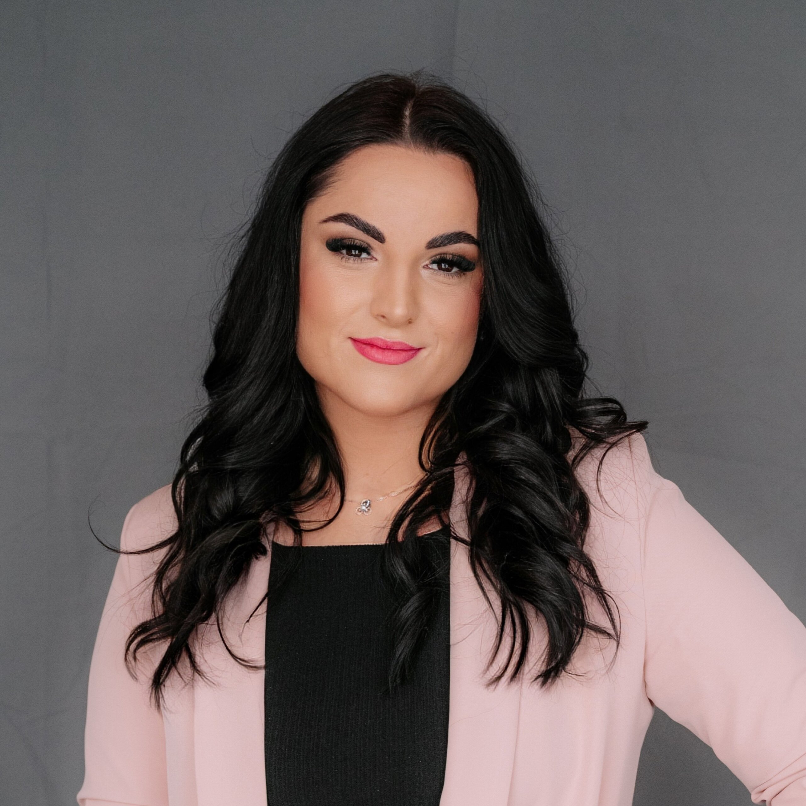 Portrait of a woman with long dark hair wearing a pink blazer over a black top, smiling at the camera against a gray backdrop.